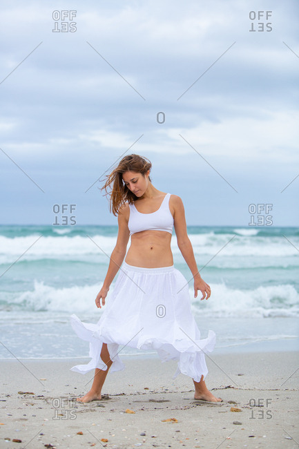 Beautiful attractive female in white outfit dancing on sand near waving sea