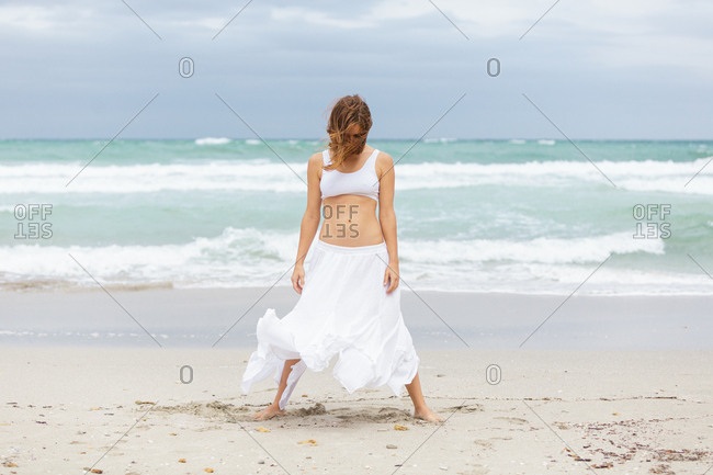 Side view of beautiful attractive female in white outfit dancing on sand near waving sea