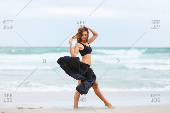 Side view of beautiful attractive female in black outfit dancing on sand near waving sea