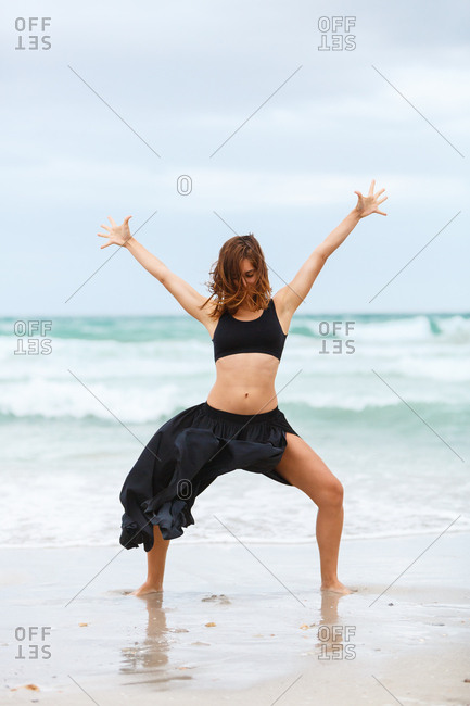 Beautiful attractive female in black outfit dancing on sand near waving sea