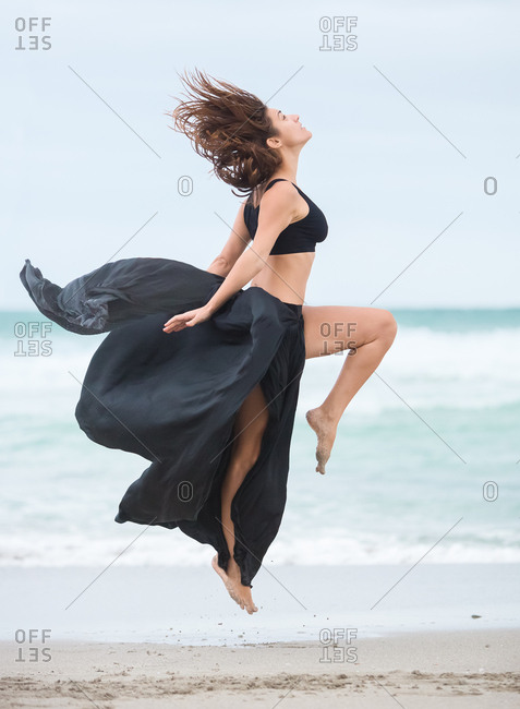 Side view of beautiful attractive female in black outfit dancing on sand near waving sea