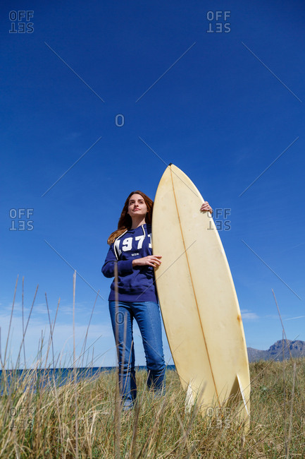Attractive long haired woman holding surfboard and standing on dry seaside hill on background of serene seascape