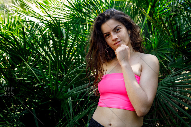 Beautiful woman wearing pink top standing in tropical green bushes on summertime and holding chin