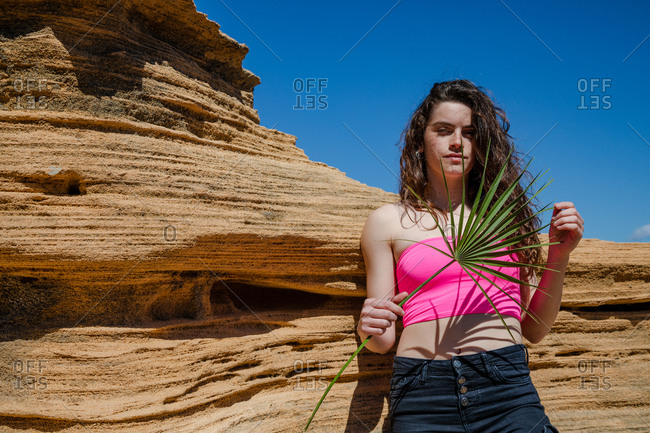 Attractive woman with green tropical leaf in hand leaning on sandstone rock and looking at camera