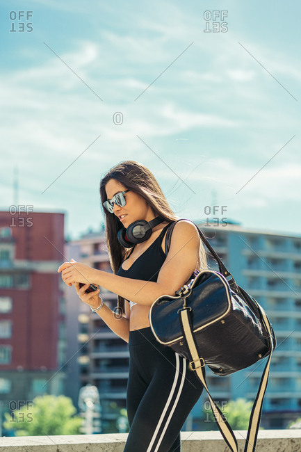 Young athletic brunette woman in sportswear sunglasses and headphones with black sport bag checking mobile phone on background of cityscape
