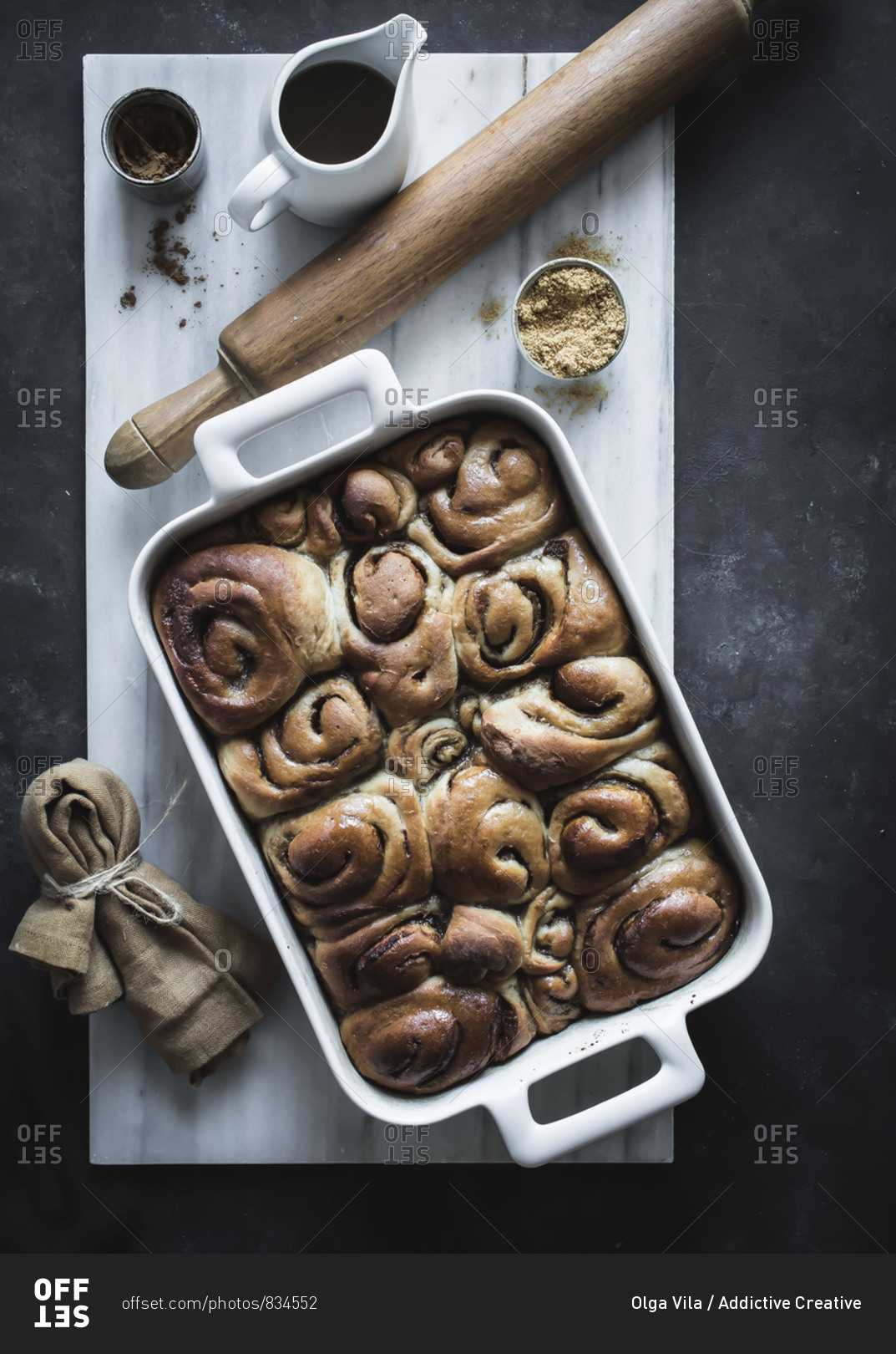 From above tray with fresh homemade cinnamon buns and wooden rolling pin on white marble board