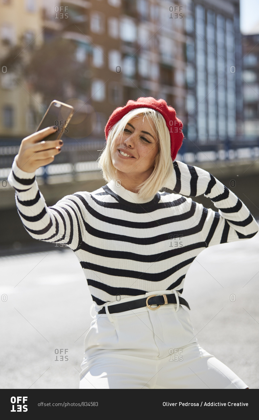 Young smiling pretty woman in French red cap, striped blouse and white shorts taking photo on urban background