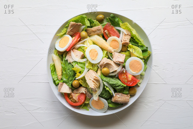From above bowl of fresh salad with lettuce, tuna, tomatoes, eggs, onion and olives on white texture background