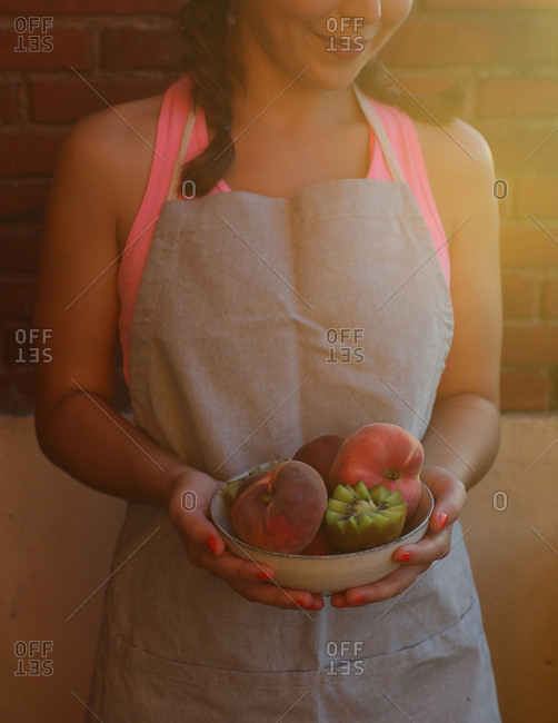 Tasty appetizing peaches and cut ripe juicy kiwi in bowl in hands of smiling woman in apron