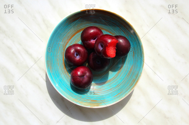 From above appetizing ripe bitten plum and whole fruit in round blue plate on marble background