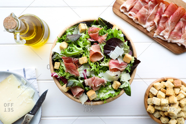 From above bowl of delicious lettuce salad with bacon and croutons placed on white tabletop near bottle of oil and slice of cheese