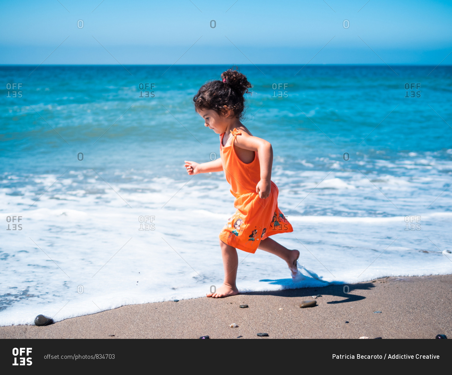 Side view of female kid in orange dress running and playing on sandy seaside and looking at sea and sky