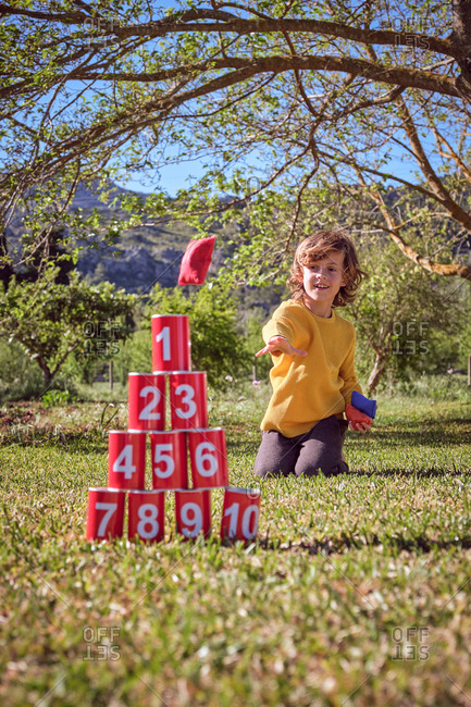 Cheerful kid kneeling and throwing balls in pyramid of red tin cans on nature background