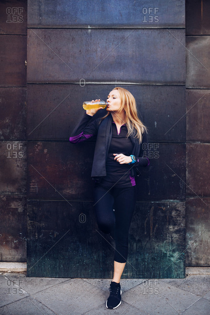 Young blonde caucasian woman drinking isotonic drink while having a rest from a running session leaning in a wall