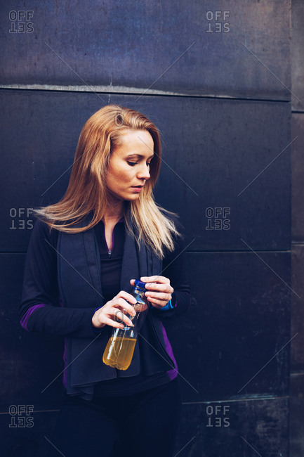 Young blonde caucasian woman drinking isotonic drink while having a rest from a running session leaning in a wall