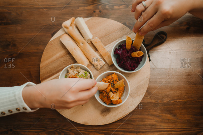 From above crop unrecognizable hands grabbing assorted vegetable dips with crispy crunchy bread in bowl on round stand on table