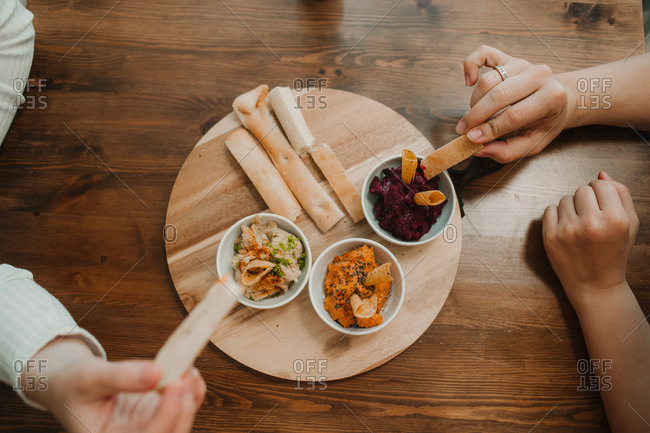 From above crop unrecognizable hands grabbing assorted vegetable dips with crispy crunchy bread in bowl on round stand on table