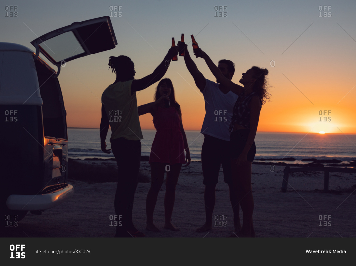 Side view of group of diverse friends toasting beer bottles near camper van during sunset