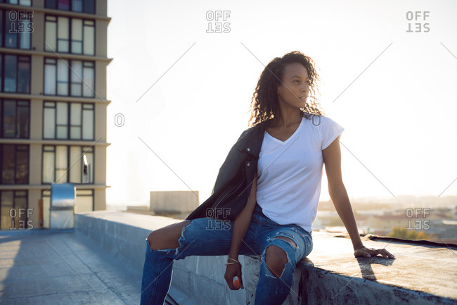 Front view of a young African-American woman with a leather jacket over shoulder sitting while looking away from the camera on a rooftop with a view of a building and sunlight