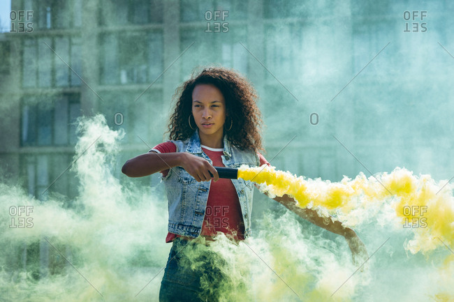 Front view of a young African-American woman wearing a denim vest holding a smoke maker producing yellow smoke on a rooftop with a view of a building and sunlight