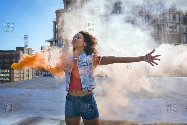 Front view of a young African-American woman wearing a denim vest with arms stretched out and holding a smoke maker producing orange smoke  on a rooftop with a view of a building and sunlight