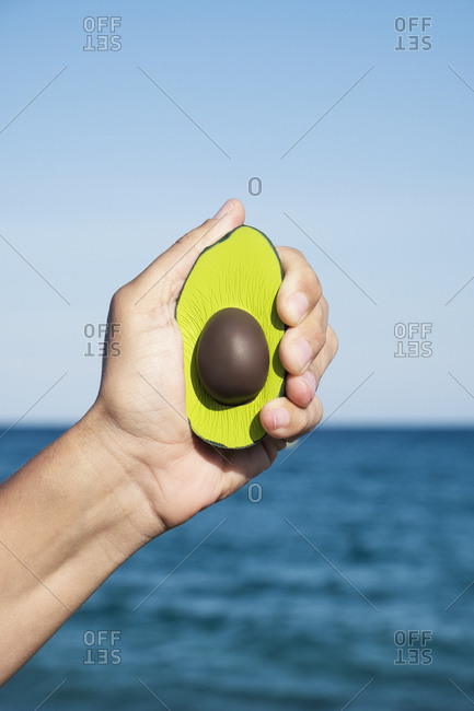 Closeup of the hand of a caucasian man squeezing a fake avocado with his hand, in front of the ocean