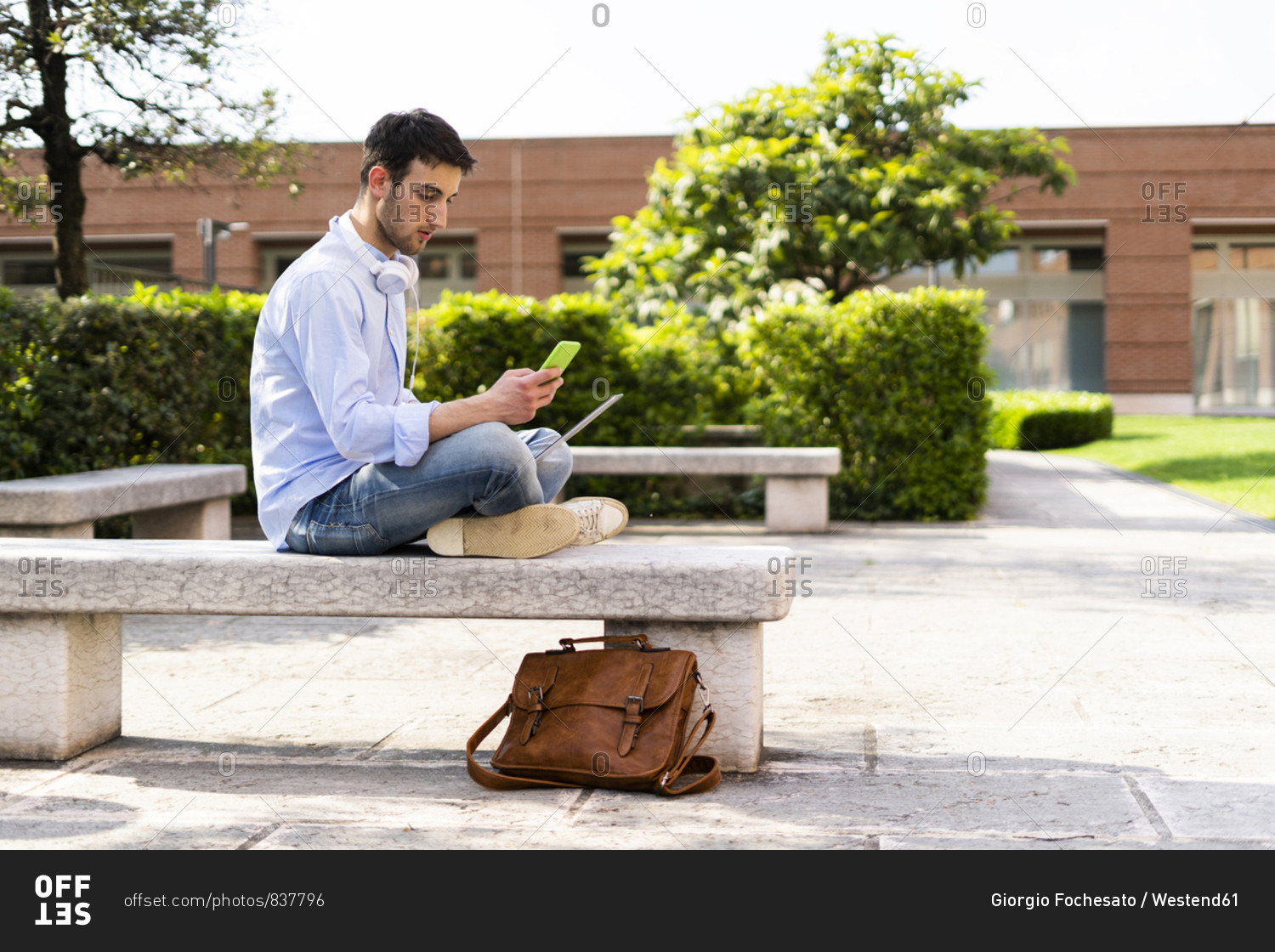 Young man using laptop and smartphone- headphones around neck- sitting on a bench