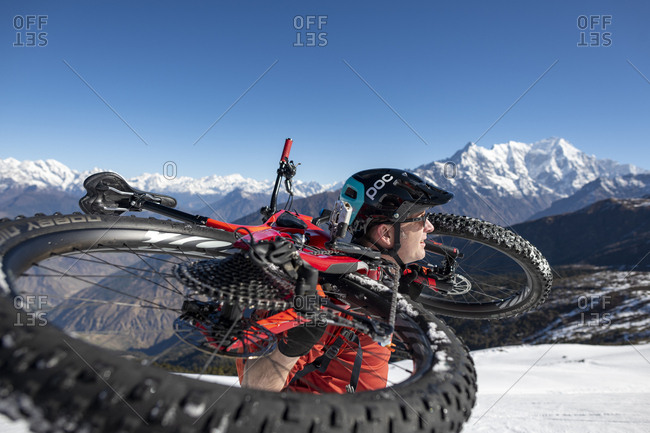Gosainkund, Nepal - March 28, 2019: A mountain biker carries his bike up a snow covered hillside in the Himalayas with views of the Langtang mountain range in the distance
