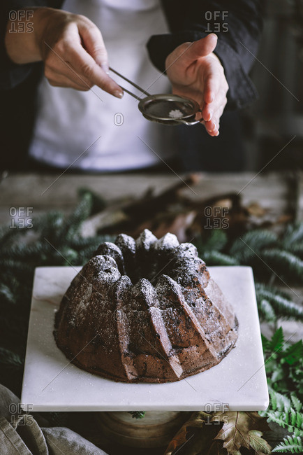 Crop person standing near table and sprinkling chocolate cake with powdered sugar