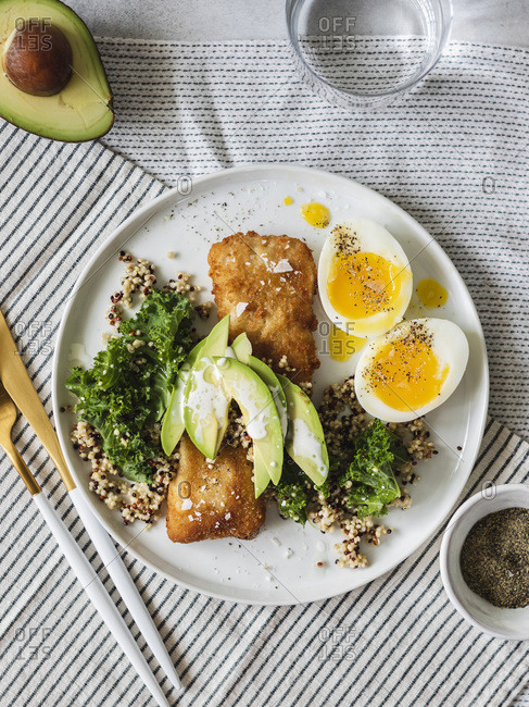 Baked fish with quinoa, kale and avocado with soft boiled eggs on the side served on white plate