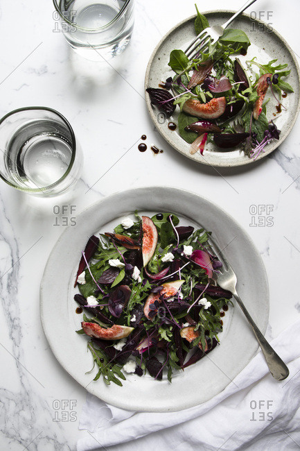 A salad of roasted beetroot, purple carrots and purple onions, with fresh figs, mixed leaves and microherbs, dotted with feta cheese. Two plated servings are on a marble tile surface with glasses of water.