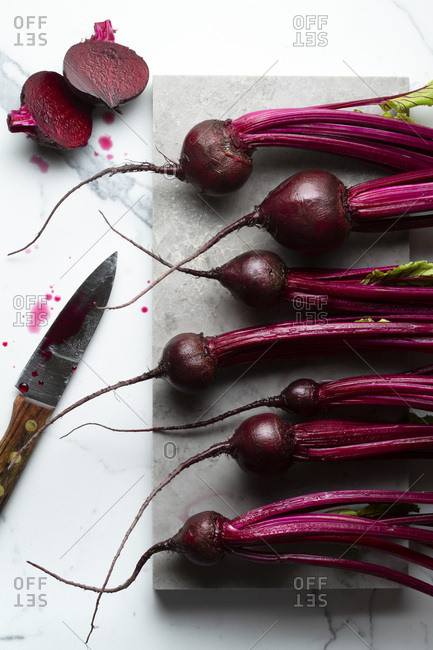 Washed baby beetroots on a small grey marble board, on a marble tile surface. A knife with beetroot juice and a cut beetroot are alongside.