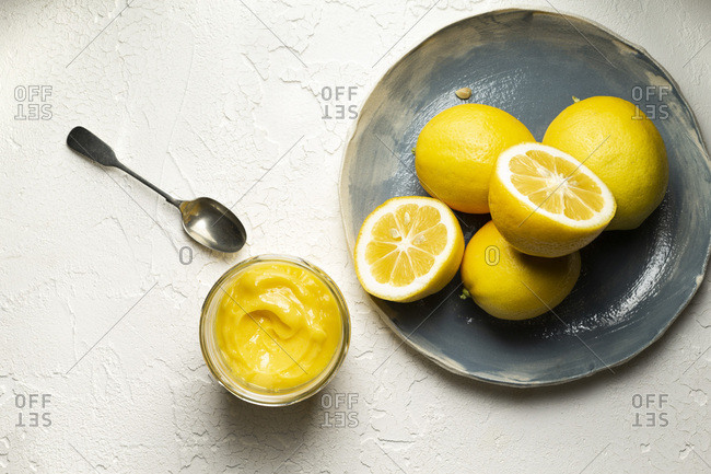 Fresh lemons, whole and sliced in half, arranged on a blue hand made plate. A jar of home made lemon curd and a spoon are alongside.