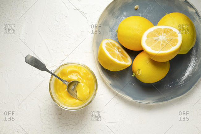 Fresh lemons, whole and sliced in half, arranged on a blue hand made plate. A spoon in jar of home made lemon curd is alongside.