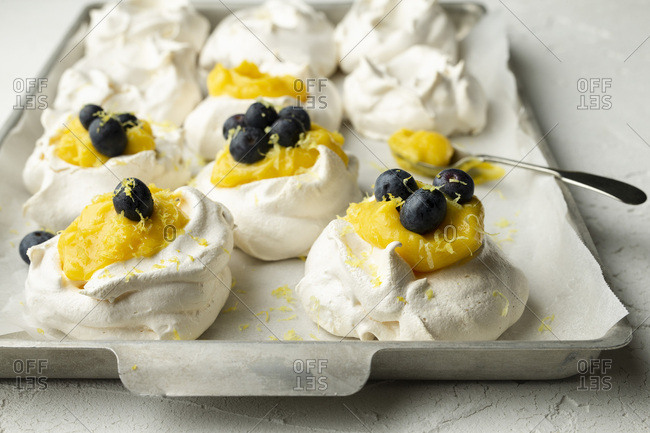 Meringues with lemon curd and blueberries on a baking tray with parchment, on a white textured background. A spoon of lemon curd is on the tray.