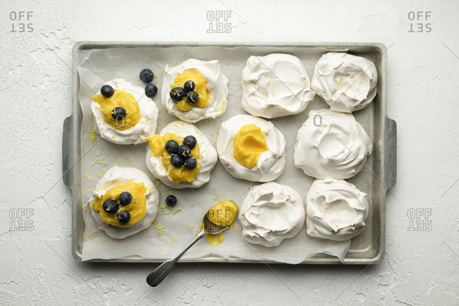 Meringues with lemon curd and blueberries on a baking tray with parchment, on a white textured background. A spoon of lemon curd is on the tray. Lemon zest has been grated over.