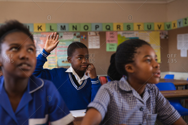 Front view of a young African schoolboy sitting at his desk, raising his hand to answer a question during a lesson in a township elementary school classroom, in the foreground two schoolgirls are listening attentively