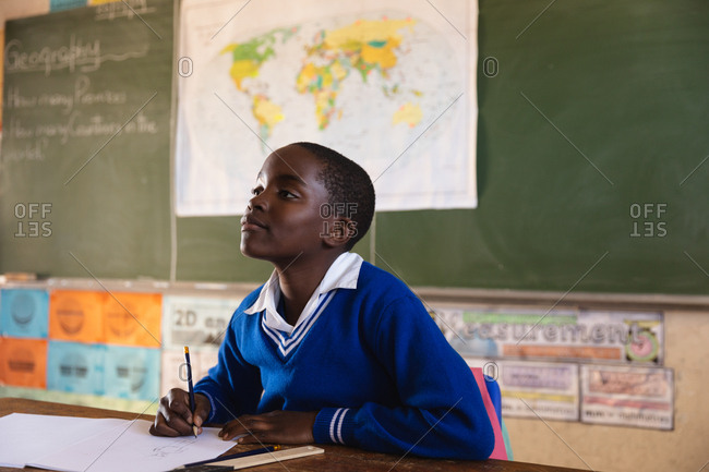 Side view close up of a young African schoolboy sitting at a desk looking up while writing in his note book and listening attentively during a lesson in a township elementary school classroom