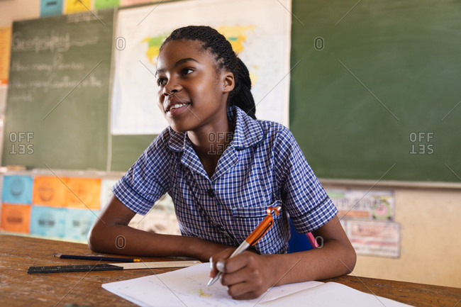 Front view close up of a young African schoolgirl sitting at a desk smiling, writing in her note book and listening attentively during a lesson in a township elementary school classroom