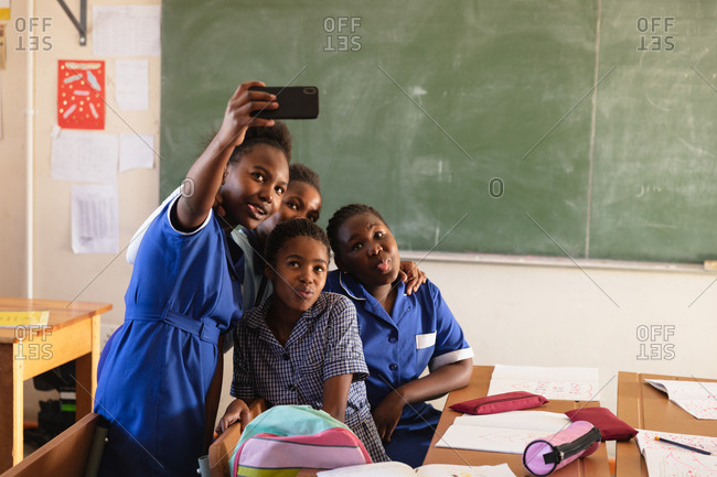 Front view close up of a group of young African schoolgirls having fun posing and taking selfies with a smartphone during a break from lessons in a township elementary school classroom