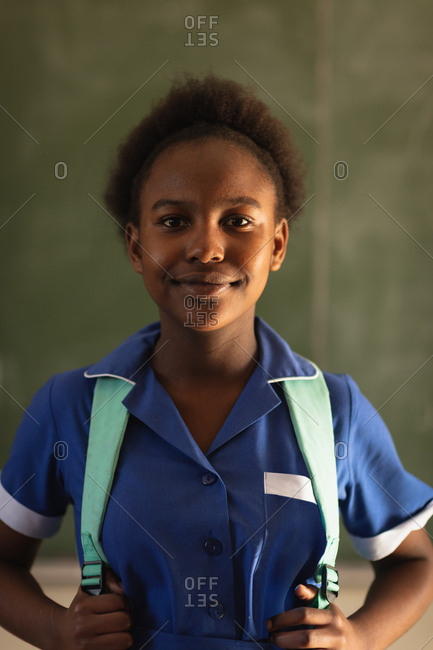 Portrait close up of a young African schoolgirl wearing her school uniform and schoolbag, looking straight to camera smiling, at a township elementary school