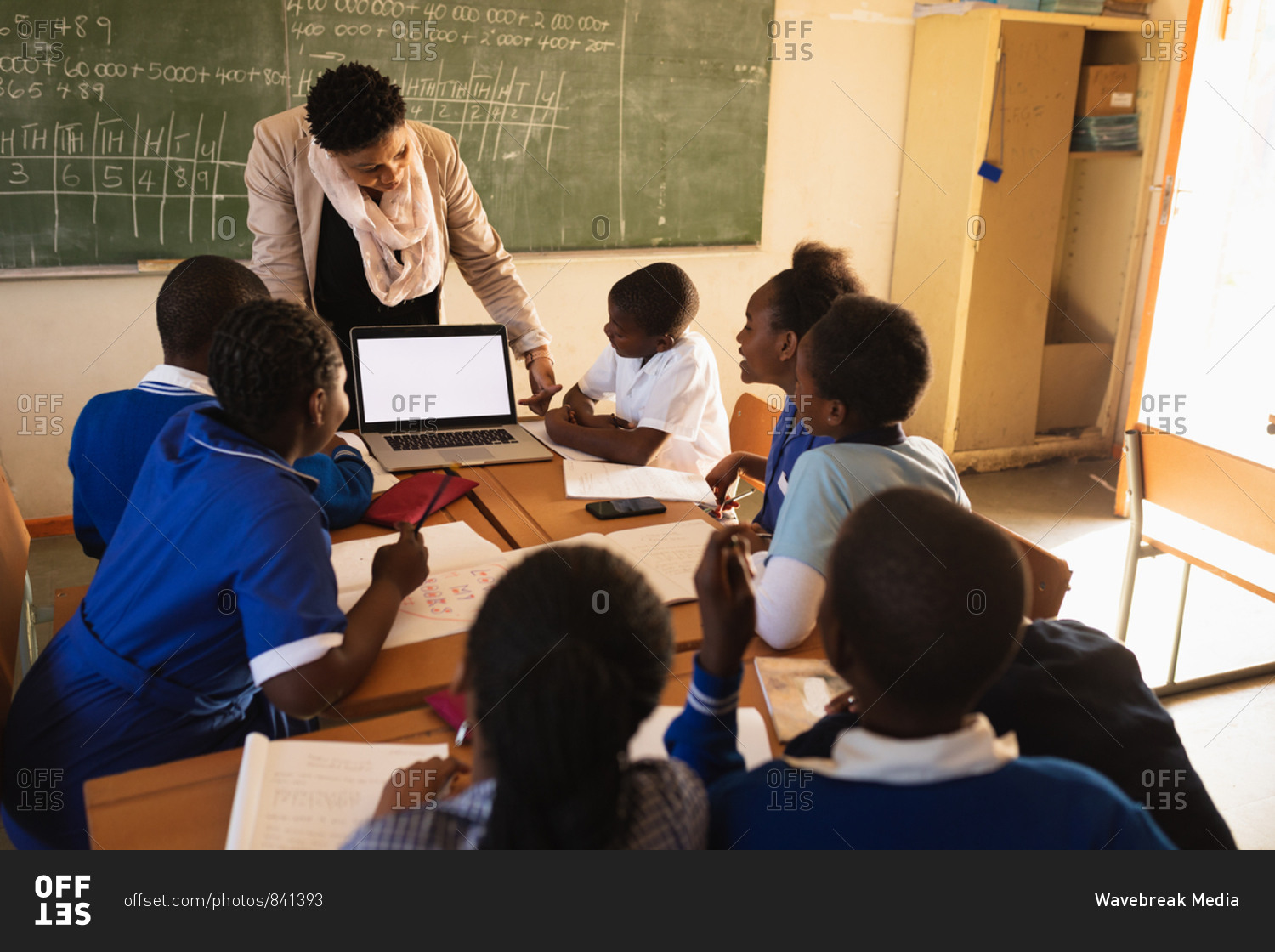 Front view of a middle aged African school teacher standing in front of the blackboard showing her pupils a laptop during a lesson in a township elementary school classroom. In the foreground her pupils are sitting at desks watching