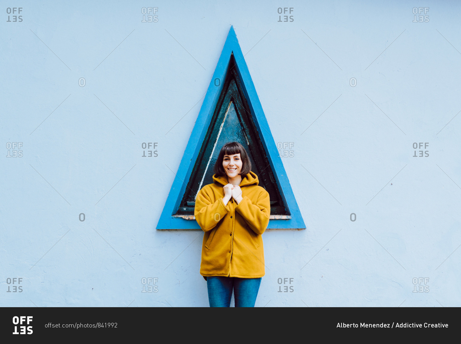 Young woman in yellow warm coat smiling and looking at camera while standing against triangle window and gray wall of building