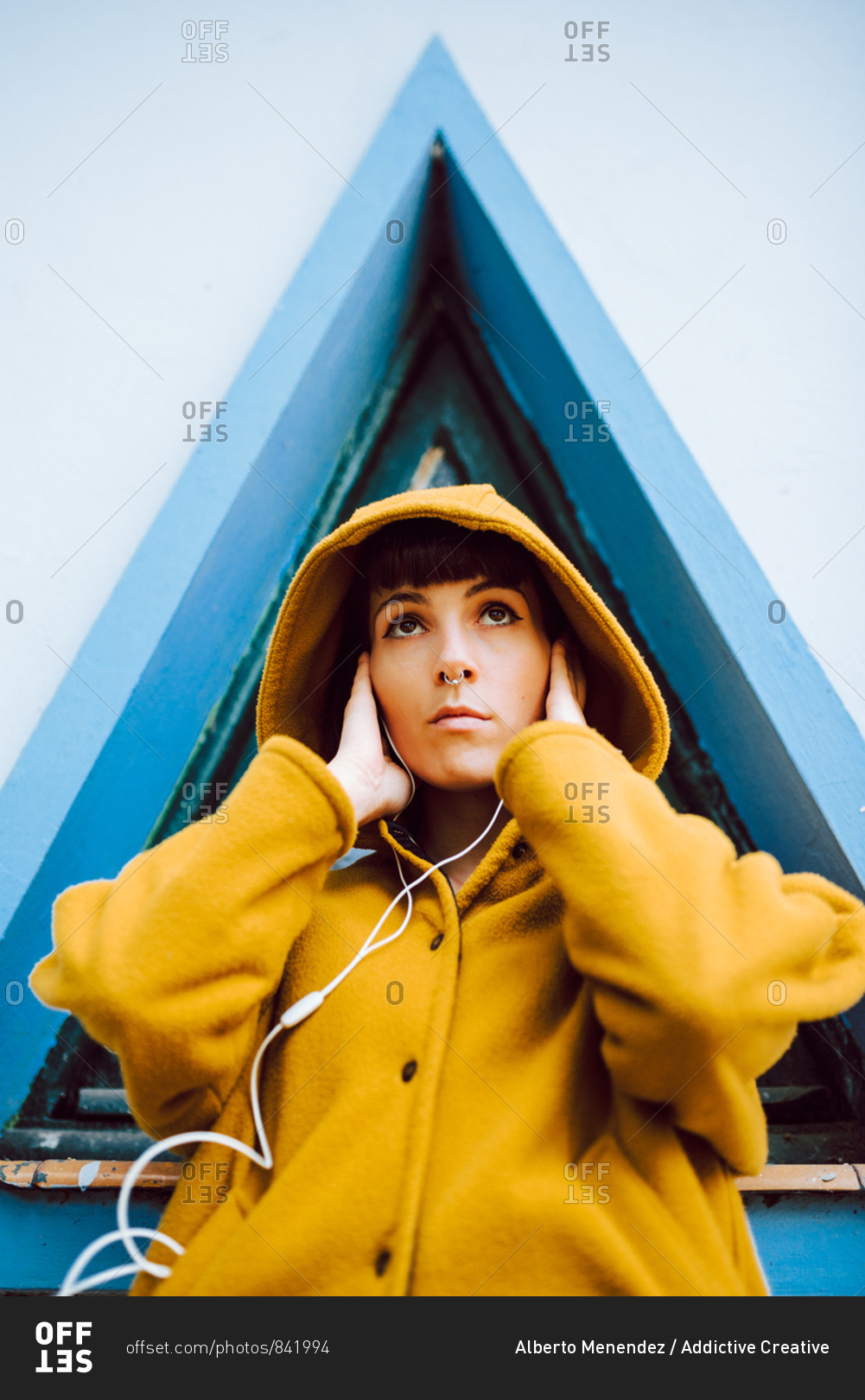 Young woman in yellow warm coat listening to the music and looking at camera while standing against triangle window and gray wall of building