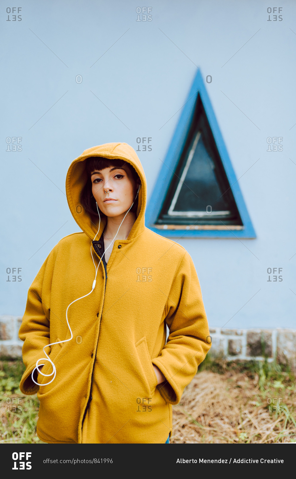 Young woman in yellow warm coat listening to the music and looking at camera while standing against triangle window and gray wall of building