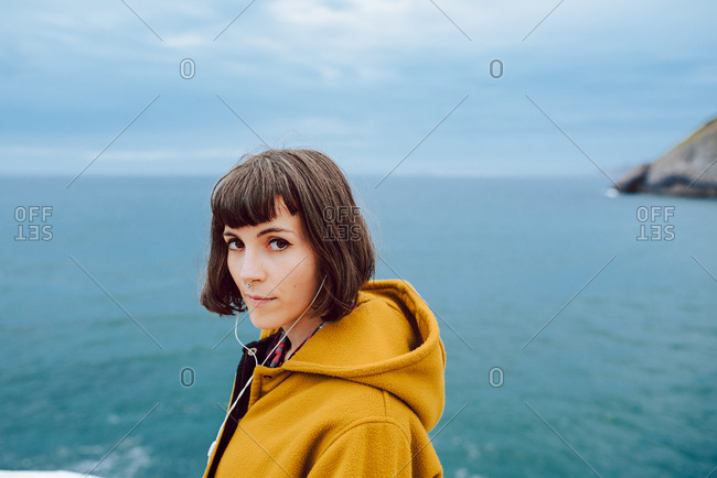 Pretty female in yellow warm jacket looking at camera while standing against rippling sea and overcast sky in nature