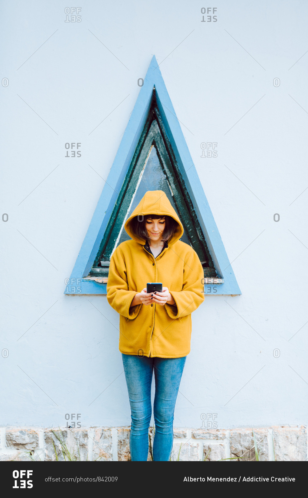 Young woman in yellow warm coat smiling and looking down while standing against triangle window and gray wall of building