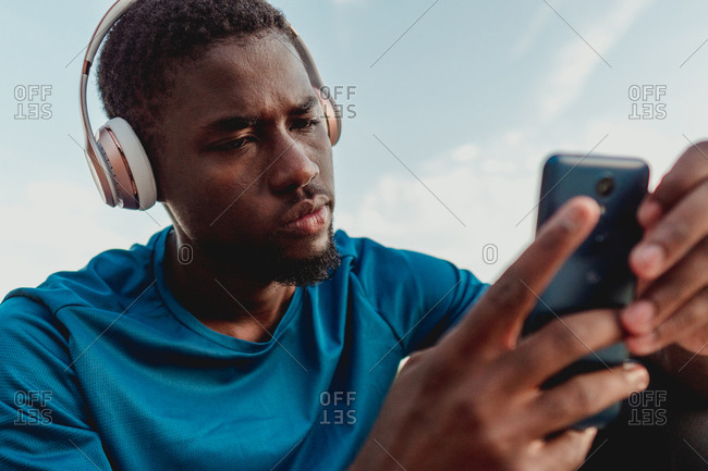 Young african american man browsing smartphone and listening to music in headphones sitting on stones in green lawn in nature