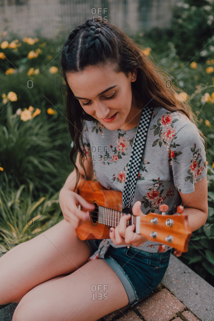 From above smiling trendy casual young woman in t-shirt playing ukulele while sitting in pavement beside flowerbed