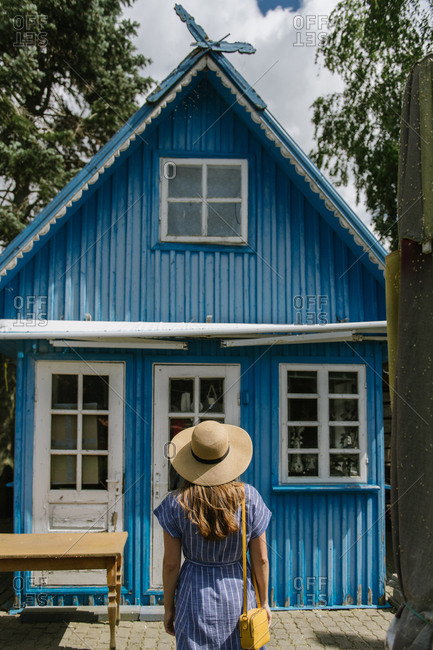 Back view of woman in straw hat and sundress standing in front of colorful blue summer house in daylight